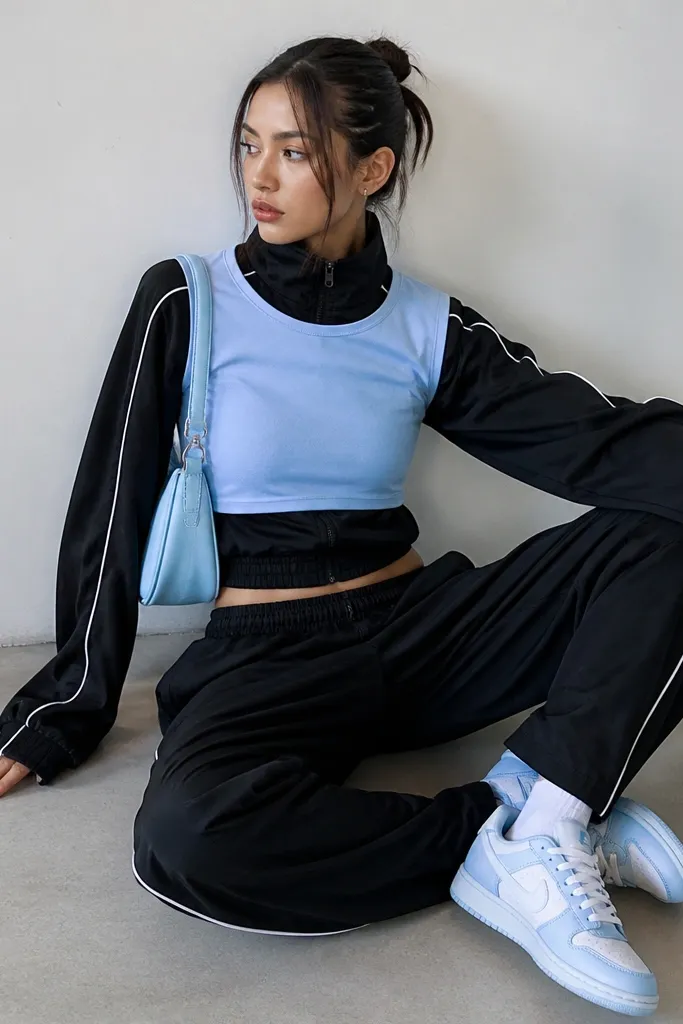 A cropped baby tee in baby blue worn over a black track jacket with white piping; matching low-top sneakers and a small shoulder bag sit at the waist.