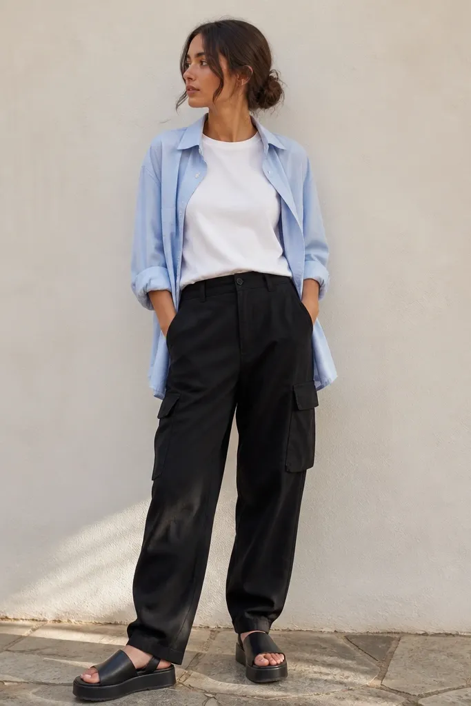 A layered summer outfit: black cargo pants with a tucked white boxy tee, an unbuttoned light blue button-up worn open over it, and platform slide sandals.