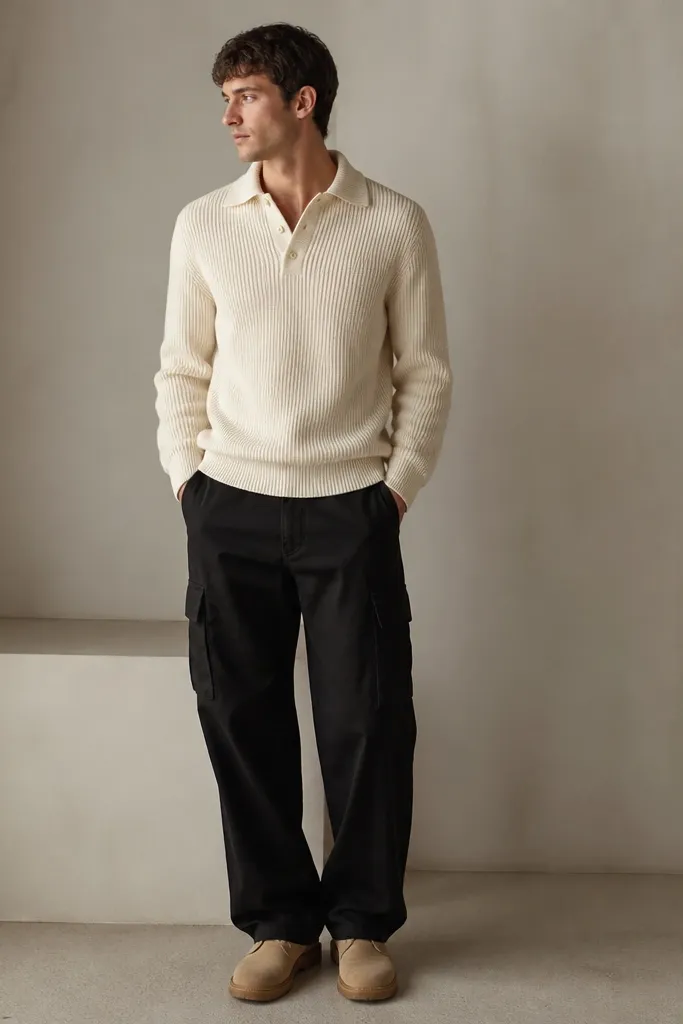 A man wearing black cargo pants with a cream knit sweater, collar slightly relaxed, sleeves down. He’s wearing tan boots or minimalist sneakers. The image shows the knit texture against the matte black fabric.