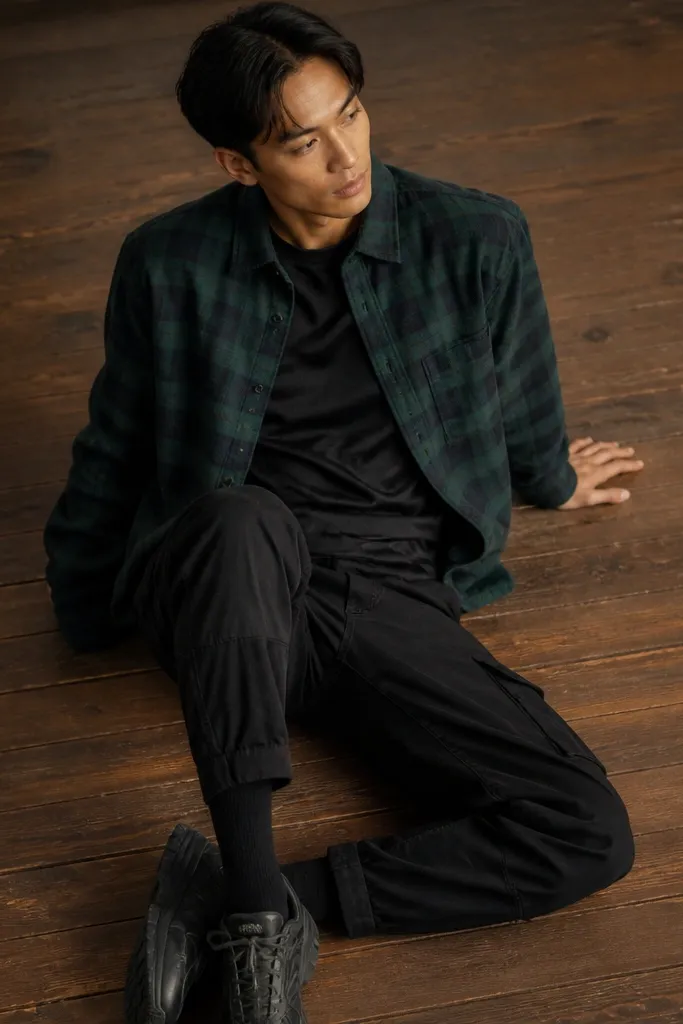 Forest green flannel shirt buttoned or worn partially open over a dark tee; black cargo pants; black crew socks visible above shoes; wooden floor background.