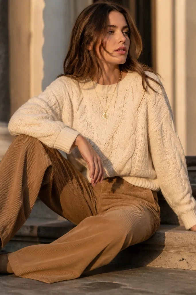 Model wearing a cream cable knit sweater with camel wide-leg corduroy pants, layered gold necklace, and tan suede ankle boots; golden-hour lighting.