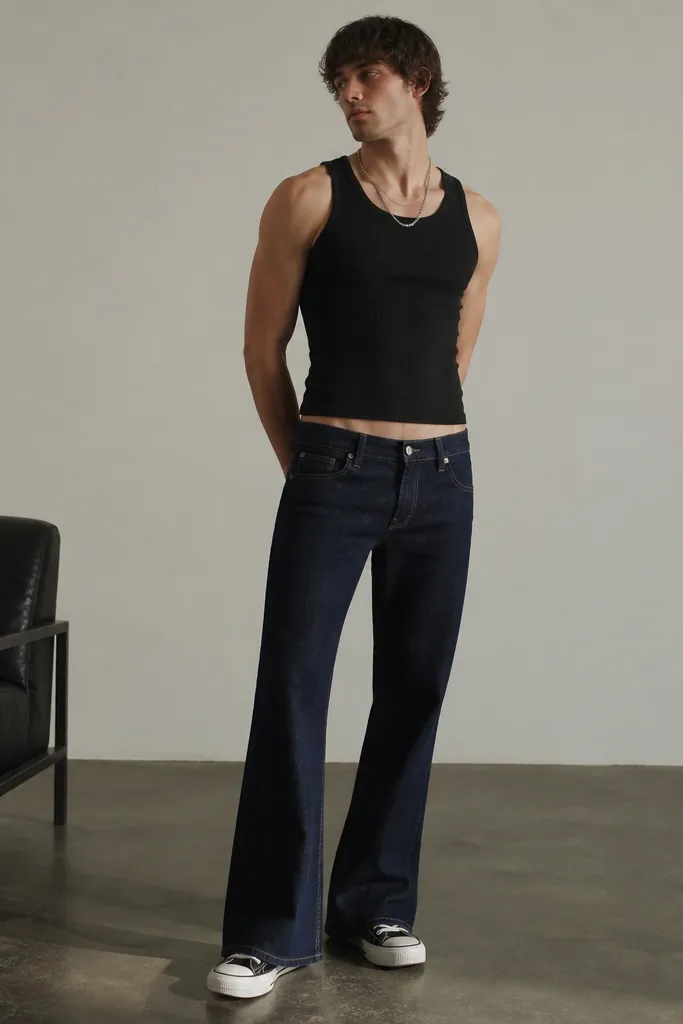 A man wearing low-rise flare jeans in dark indigo with a fitted ribbed tank underneath. The waistband sits slightly lower on the hips. He wears platform-style sneakers and a silver chain that catches the light.