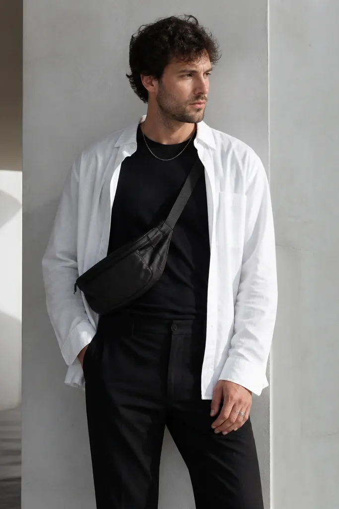 A man in an oversized white button-up worn open over a fitted black tee. A small belt bag sits across the chest/waist, and the man wears straight-leg black pants. Accessories include a thin chain and simple rings.