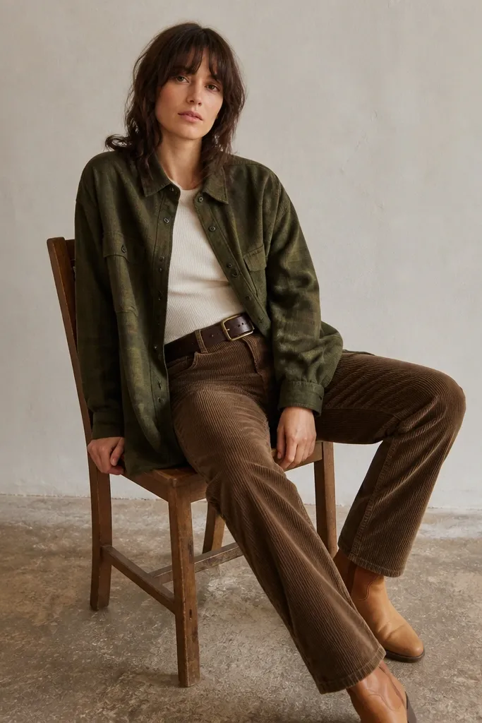 Olive green oversized flannel button-up layered over a fitted cream knit tee. Brown corduroy pants and a wide workwear belt are visible, with tan leather boots.