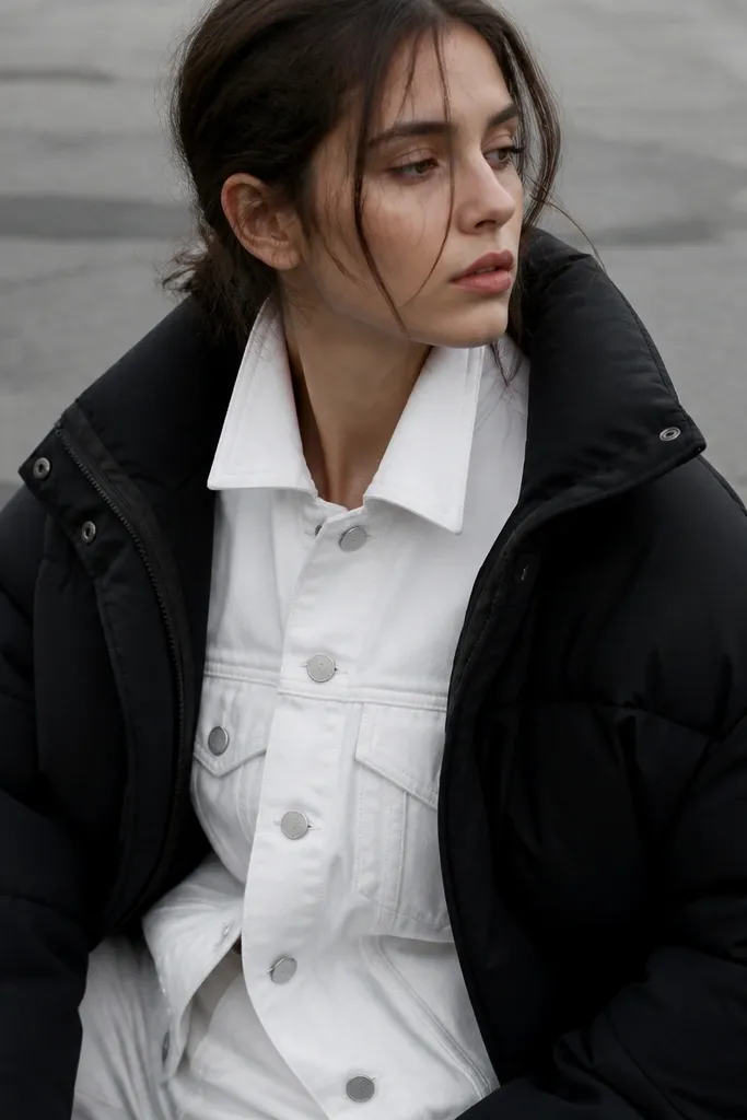 A woman wearing a bright white denim jacket under a bulky puffer; the denim looks stiff and contrasts harshly, with the outfit photographed outdoors against gray pavement.