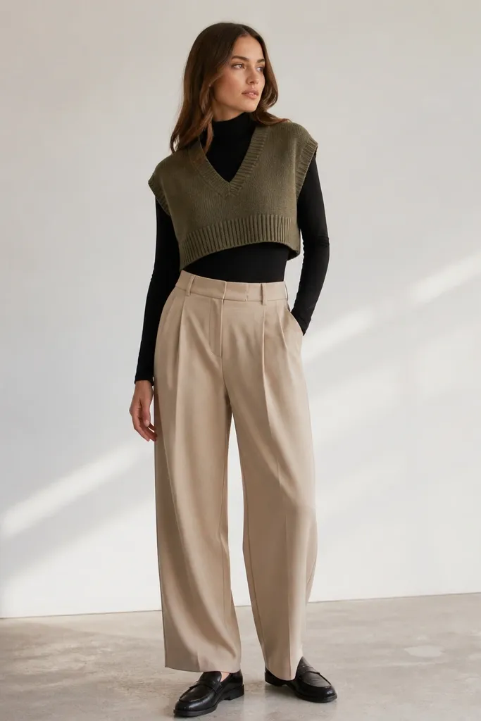 A clean studio photo: a cropped olive sweater vest over a black mock-neck bodysuit, styled with high-waist beige trousers and sleek black loafers.