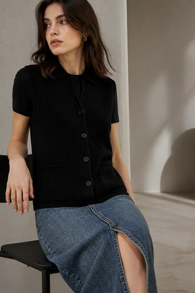 A black knit button-up sweater vest with a subtle zip-front or concealed placket detail. It’s worn over a fitted black tee and paired with a mid-length denim skirt. The vest’s storage pocket area is visible as a clean seam on the lower side.