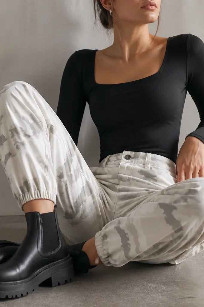 Close-up of white camo pants paired with a fitted black bodysuit, chunky black Chelsea boots, and small silver hoop earrings; the camera angle shows the ankles and waistband clearly.
